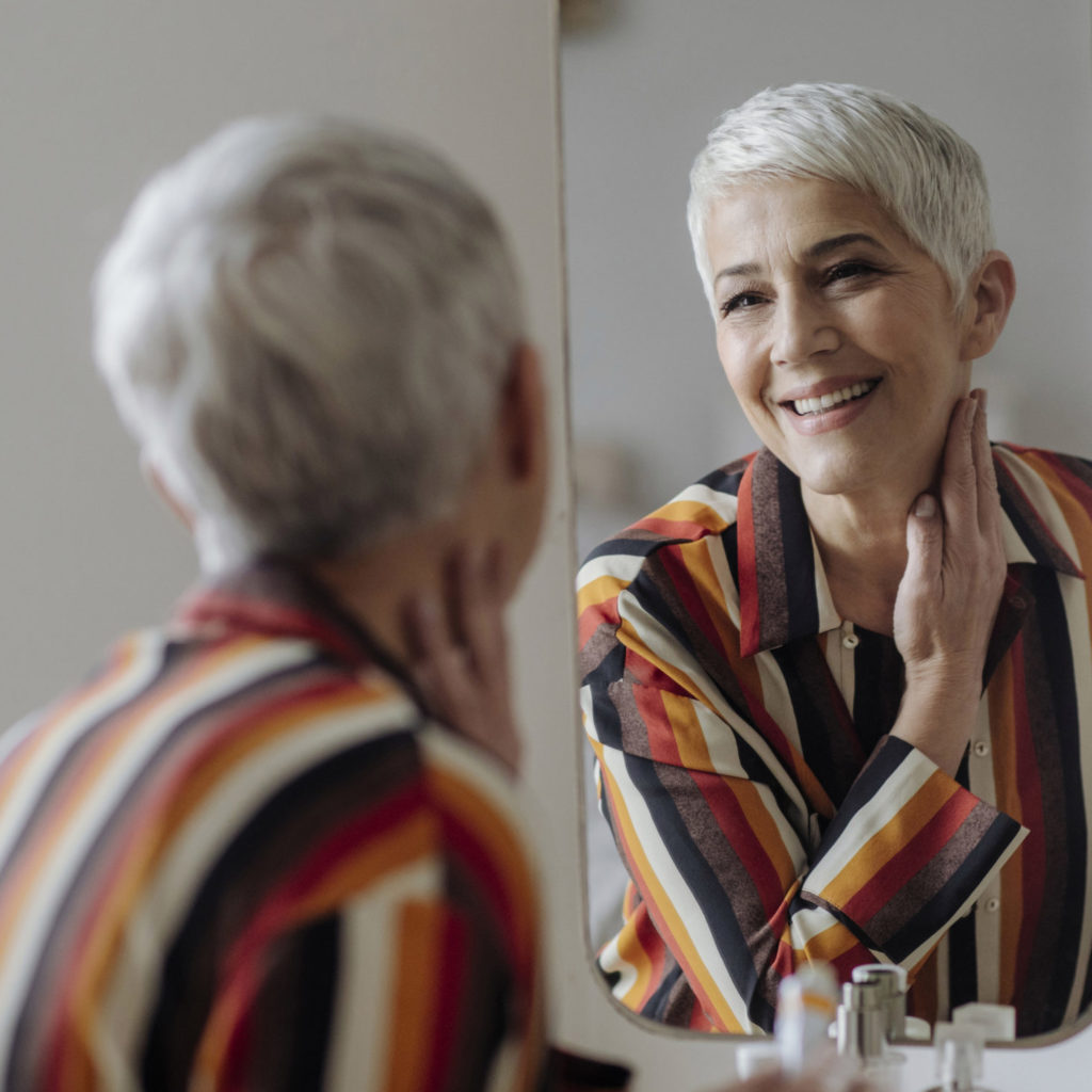 Beautiful smiling Caucasian grey-haired smiling woman looking herself in the mirror and applying face cream.