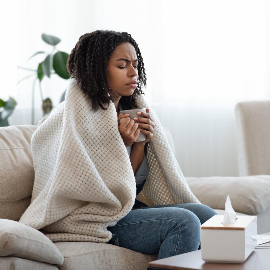 Sick Black Woman Suffering From Flu At Home, Drinking Tea And Resting On Couch Covered In Blanket