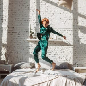 cheerful young woman jumping on bed in silk pajamas and throwing pillows up, energized.