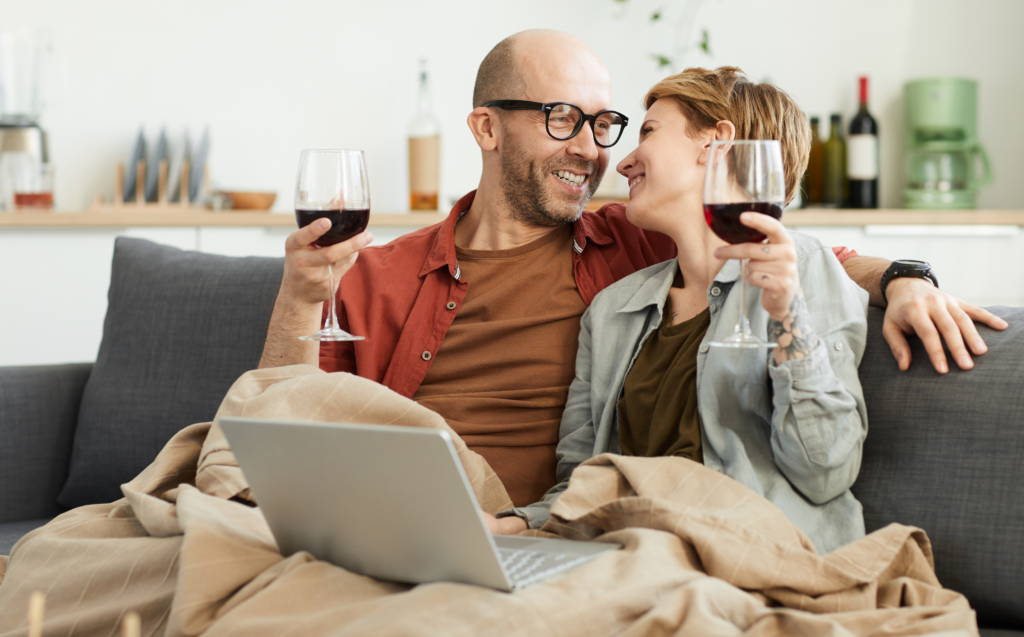 Happy mature couple sitting on sofa with glasses of red wine using laptop computer and laughing