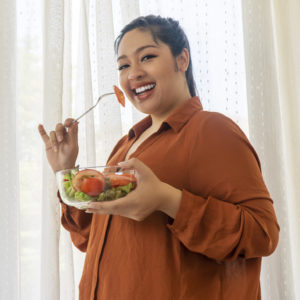 Concept food good healthy for weight loss. Smile obesity plus size women eat green organic vegetables while standing beside window in the room. Fat female holding glass bowl with salad vegetarian at home.