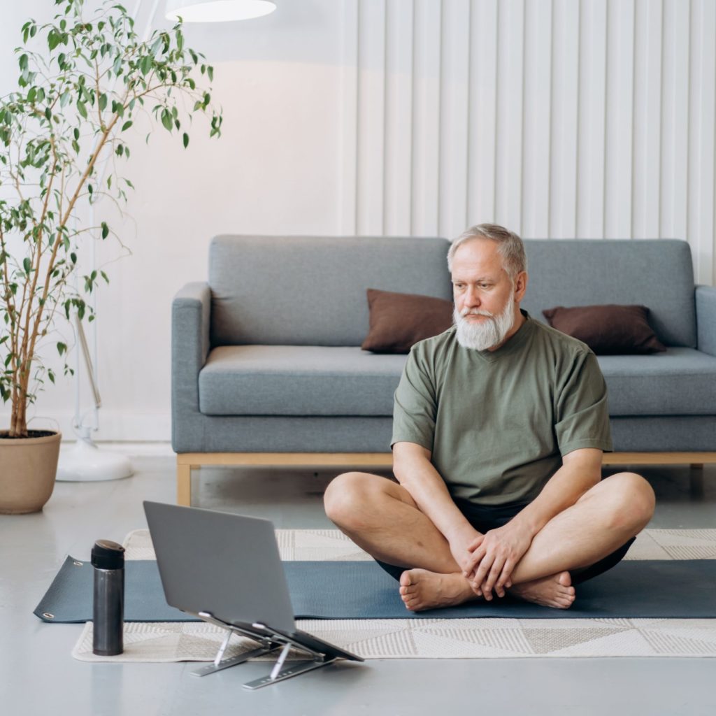 Senior man sitting on yoga mat watching a tutorial on laptop