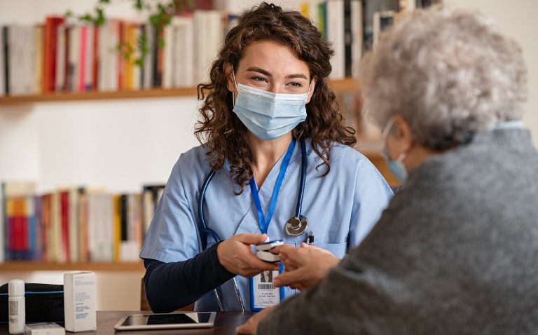 Doctor wearing surgical mask while visiting a patient at home. Senior woman sitting with doctor while doing coronavirus test and screening using oximeter.