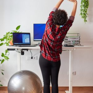 Woman stretching and working at a standing desk to avoid dangers of sitting all day.