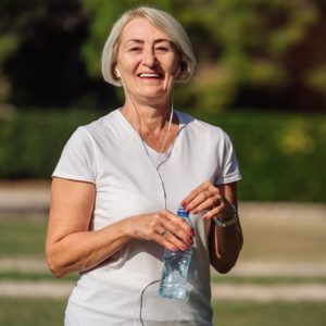 mature woman wearing headphones and drinking water outside.
