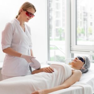 Skilled Dermatologist Wearing Special Protective Glasses and performing a procedure on patient laying on exam table.