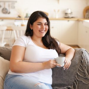 Smiling woman sitting on couch with a cup of coffee, feeling better after achieving a healthier gut.