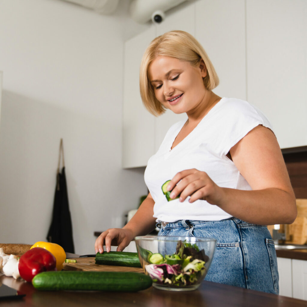 Young caucasian plump plus size woman cooking making salad, healthy food, dieting, counting calories, preparing dinner lunch at home kitchen