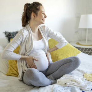 Pregnant Woman Sitting On Bed And Prepare Clothes For New Baby And Smile