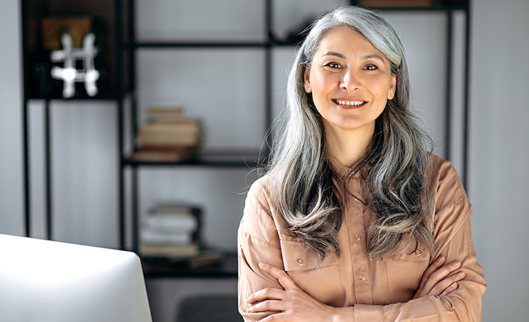 Portrait of a successful confident mature gray-haired lady, business woman, ceo or business tutor, standing in the office with arms crossed, looking and friendly smiling into the camera