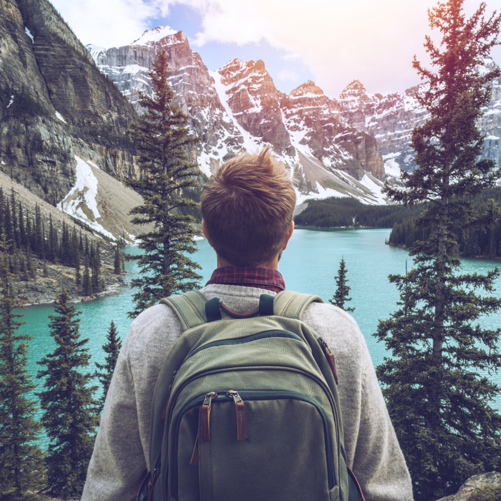 Backpacking person staring out over a scenic lake, surrounded by snowy mountain peaks.