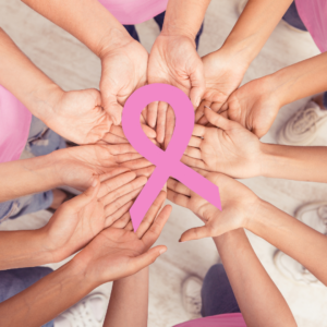 several hands placed together, holding a pink Breast Cancer Awareness ribbon.